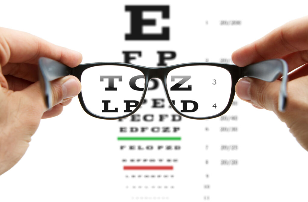 Two hands holding out a pair of glasses in from of an eye exam (Snellen) chart.