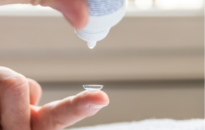 A person holding a contact lens on their figure tip while these use a bottle of solution to clean the lens