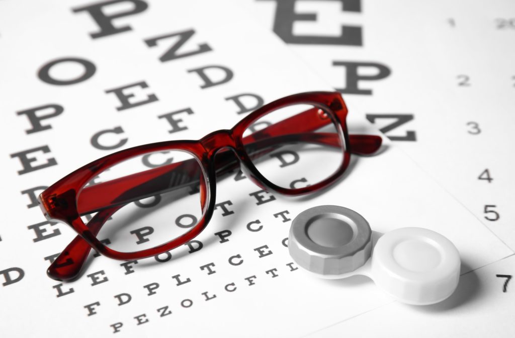 A pair of glasses, next to contact lenses in their case both resting on top of a Snellen Eye Chart