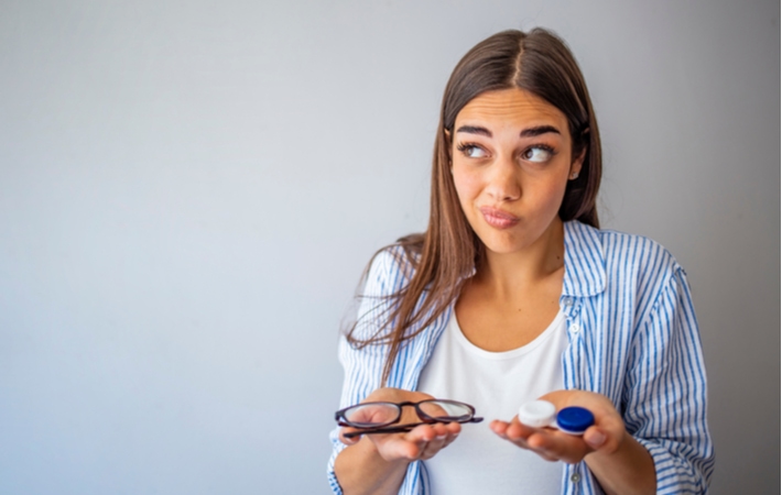 A woman pondering while she holds a pair of glasses in one hand and a pair of contact lenses in the other