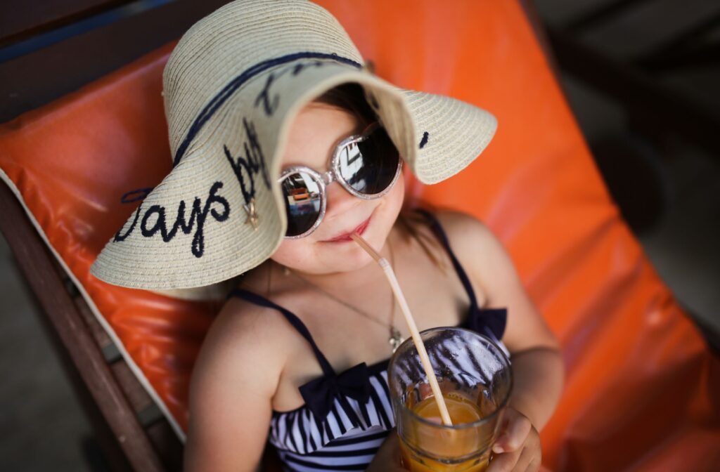 A young Girl sitting on an orange lounge chair wearing a wide-brimmed hat and sunglasses drinking through a straw.