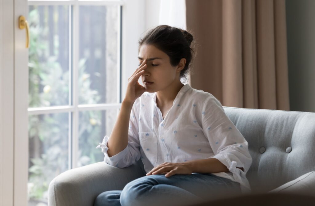 A woman sitting on the couch by a window, rubbing her eye in discomfort due to a sinus infection.