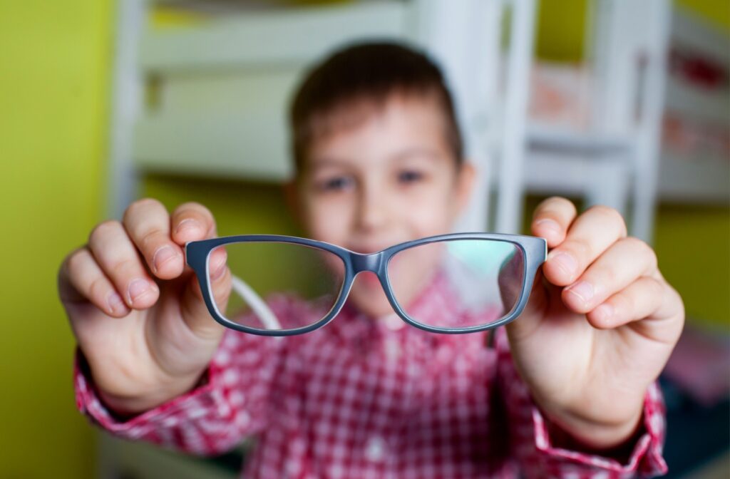 An out-of-focus young boy smiling while holding up an in-focus pair of black myopia control eyeglasses.