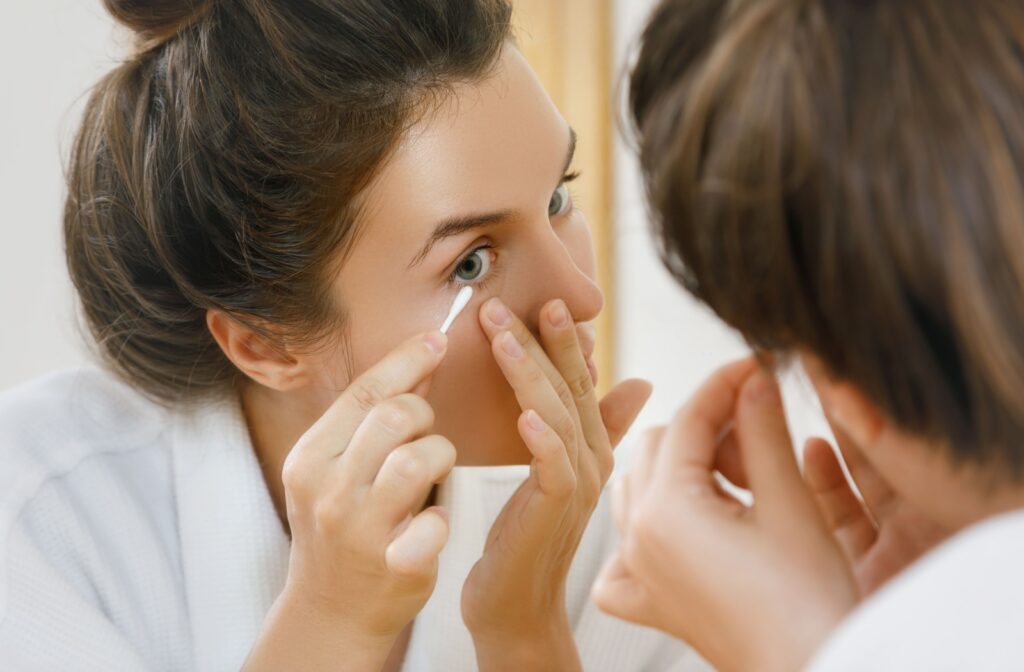 A woman looking in the mirror, using a cotton swab to clean her bottom eyelid to maintain good eyelid hygiene and prevent meibomian gland dysfunction.