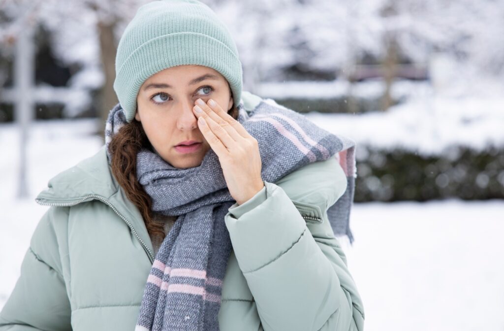 A woman standing outside in the winter, rubbing her eye due to irritation caused by the cold weather.