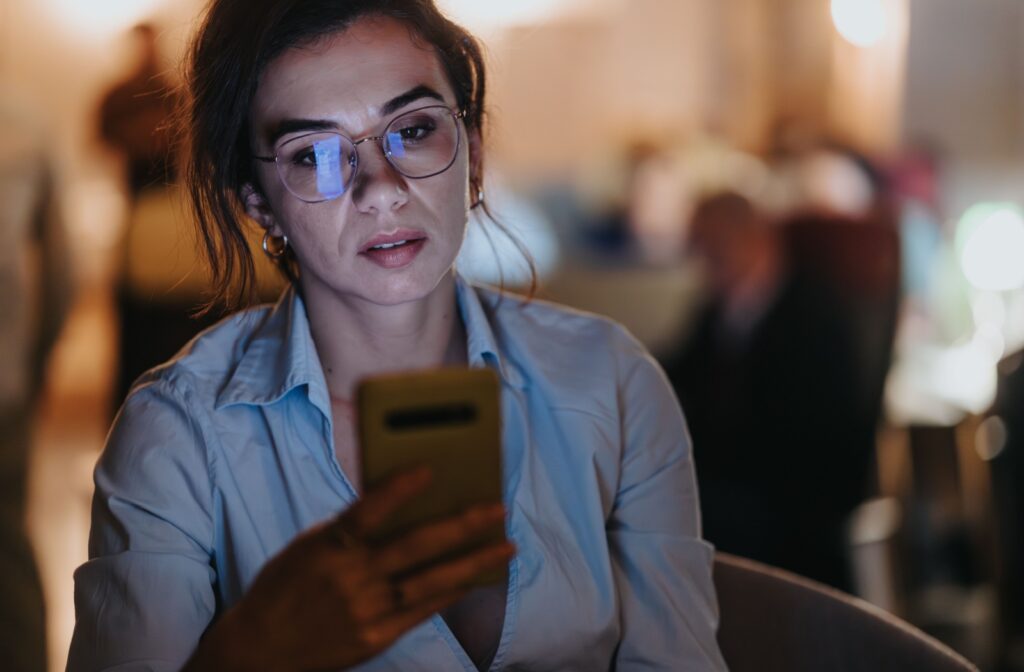 A person wearing glasses and looking at their phone in a dimly lit room
