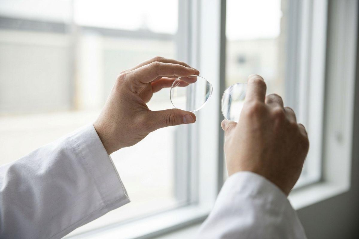 Hands in a white coat holding two clear optical lenses up toward a bright window for inspection.